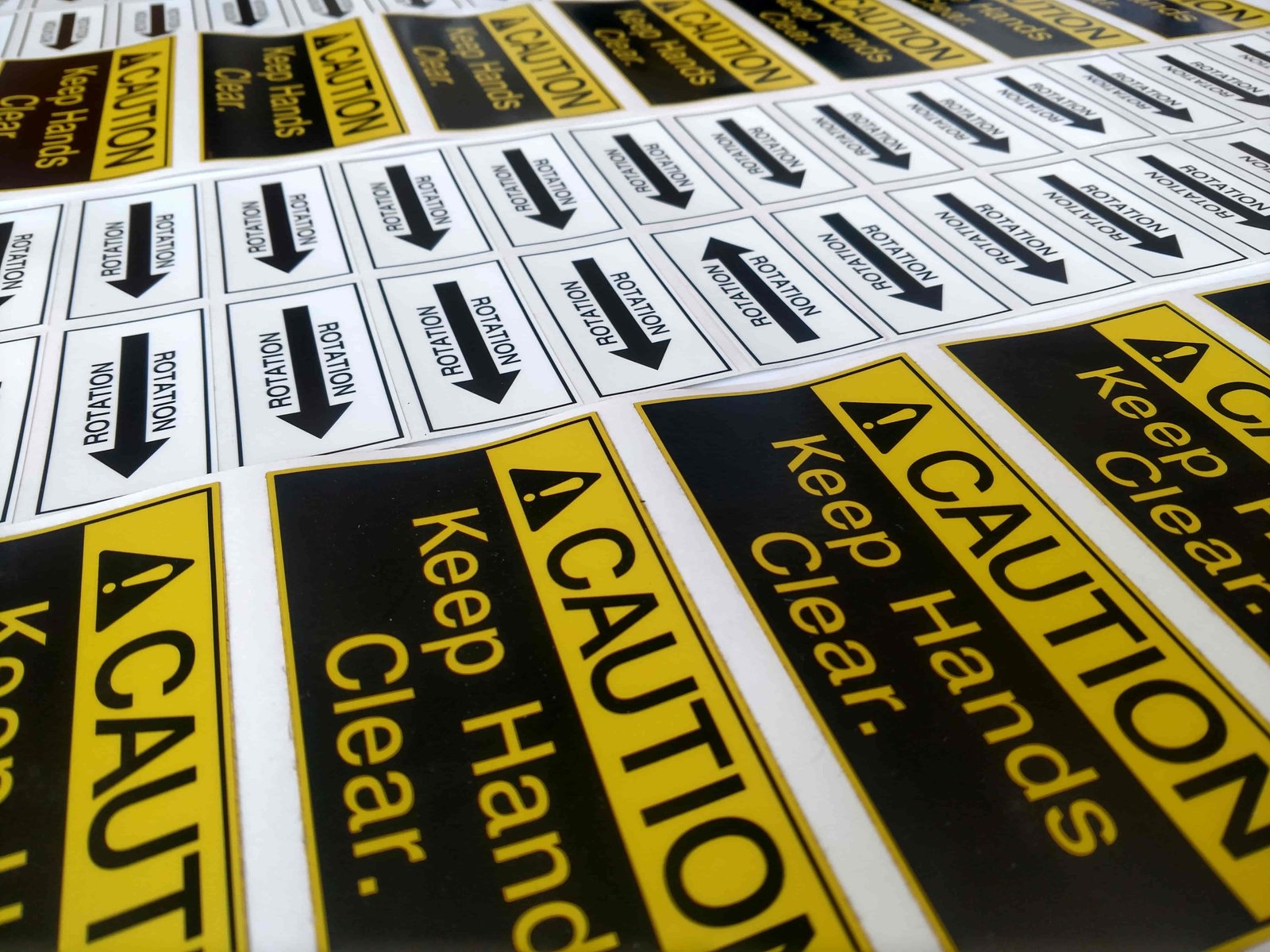 Several alternating rows of rectangular machine decals. One row is white an arrow and black text that says rotation. The other is black and yellow and says caution keep hands clear. 